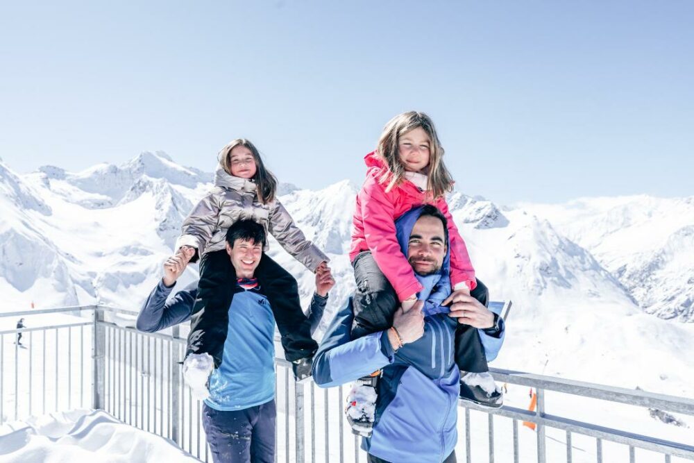 2 hommes sur un balcon portant sur leurs épaules 2 fillette avec la montagne enneigée derrière eux