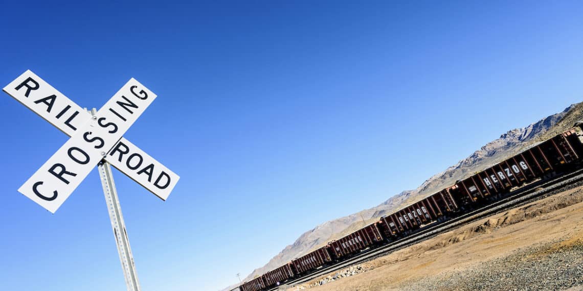 Un panneau en croix et derrièer un long train de wagon dans le désert sous un ciel bleu